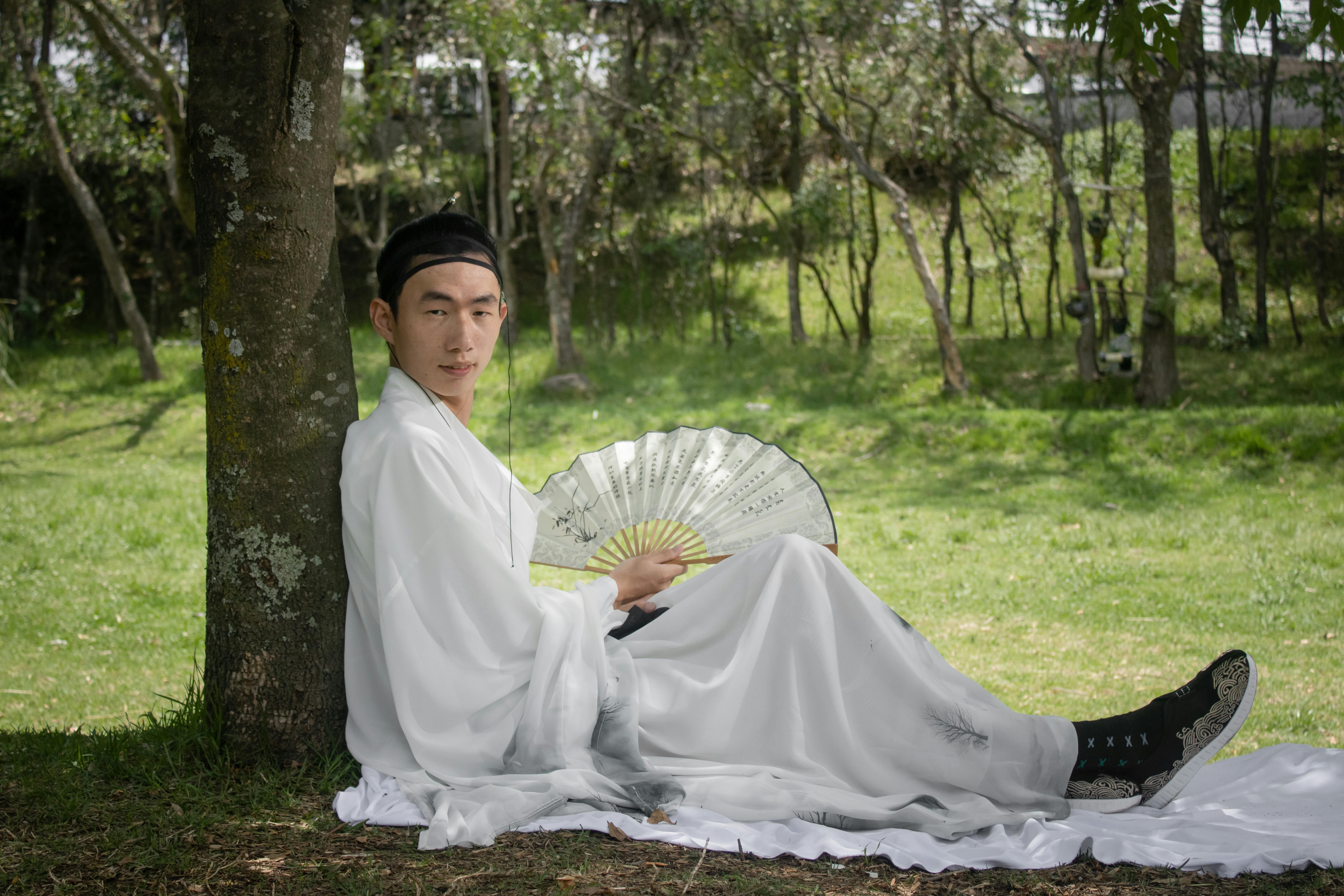 grátis Homem com túnica branca tradicional e leque relaxando ao ar livre sob uma árvore. Foto profissional