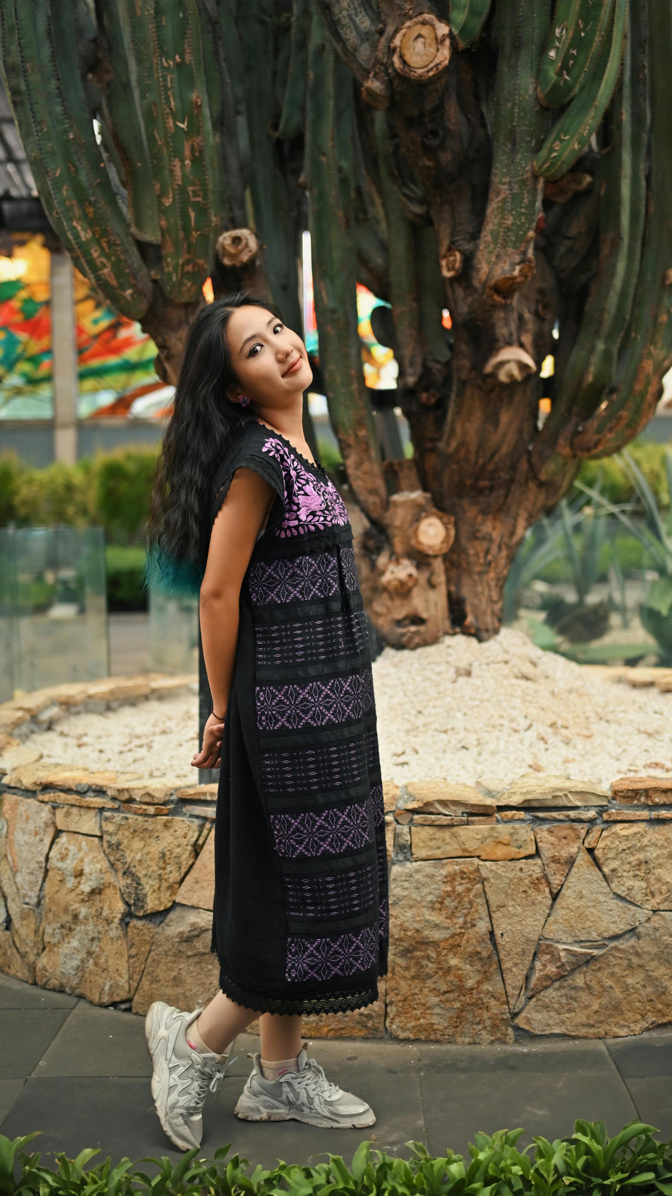 grátis Mulher com vestido bordado posa alegremente ao lado de um grande cacto ao ar livre. Foto profissional