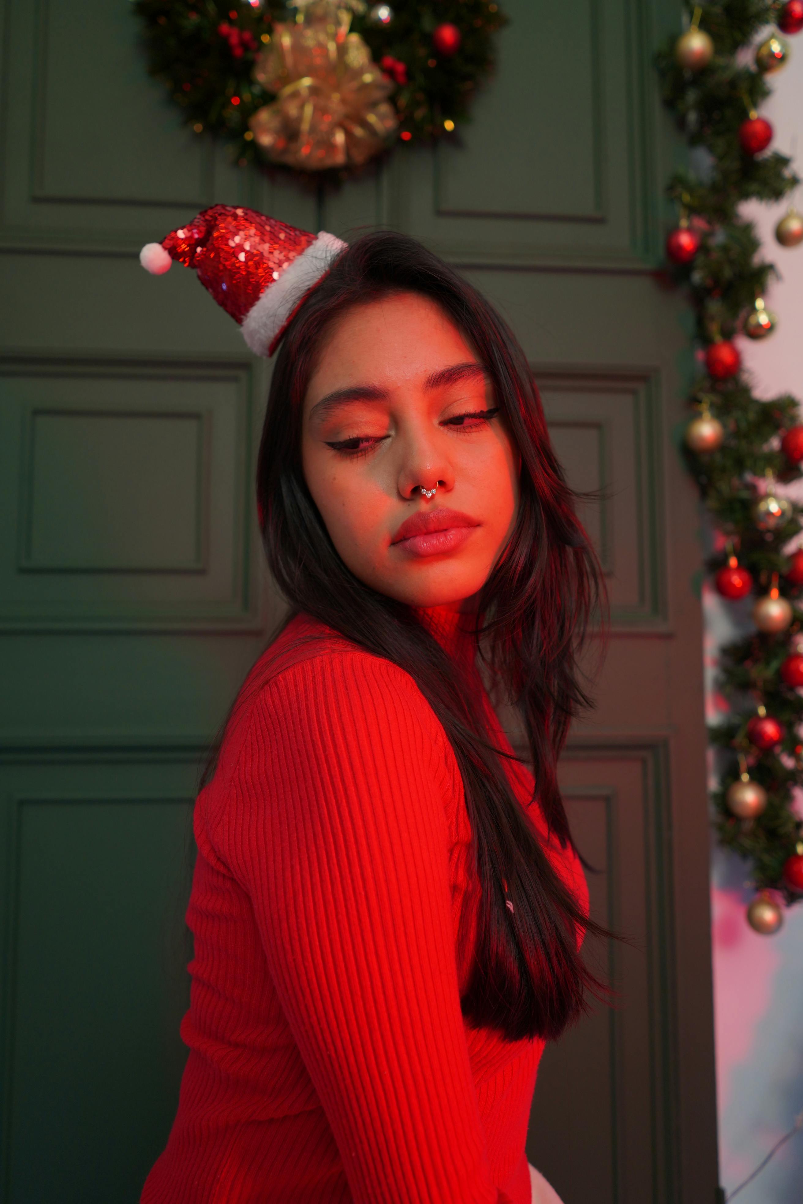 grátis Mulher com gorro de Papai Noel posando em ambiente interno com guirlandas e decorações natalinas. Foto profissional