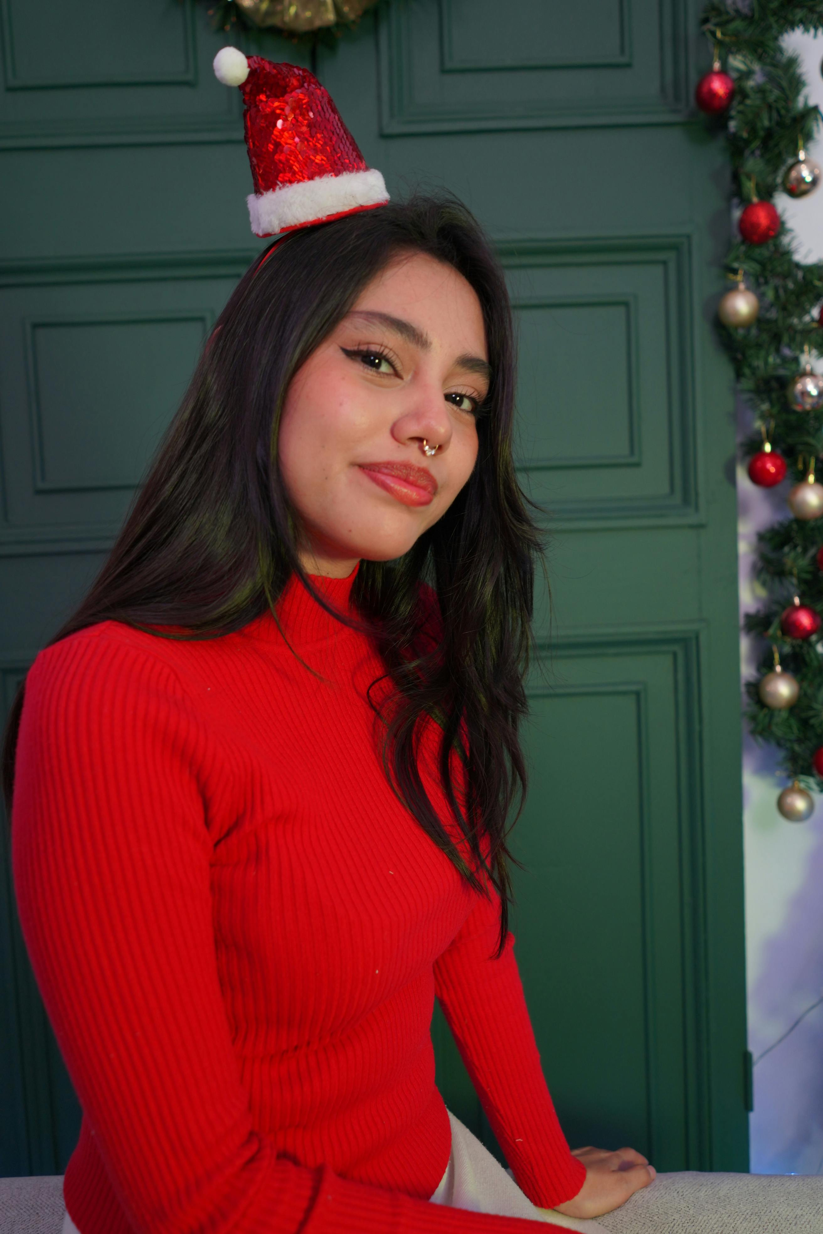 grátis Mulher alegre com suéter vermelho e gorro de Papai Noel posando em ambiente interno durante a época natalina. Foto profissional