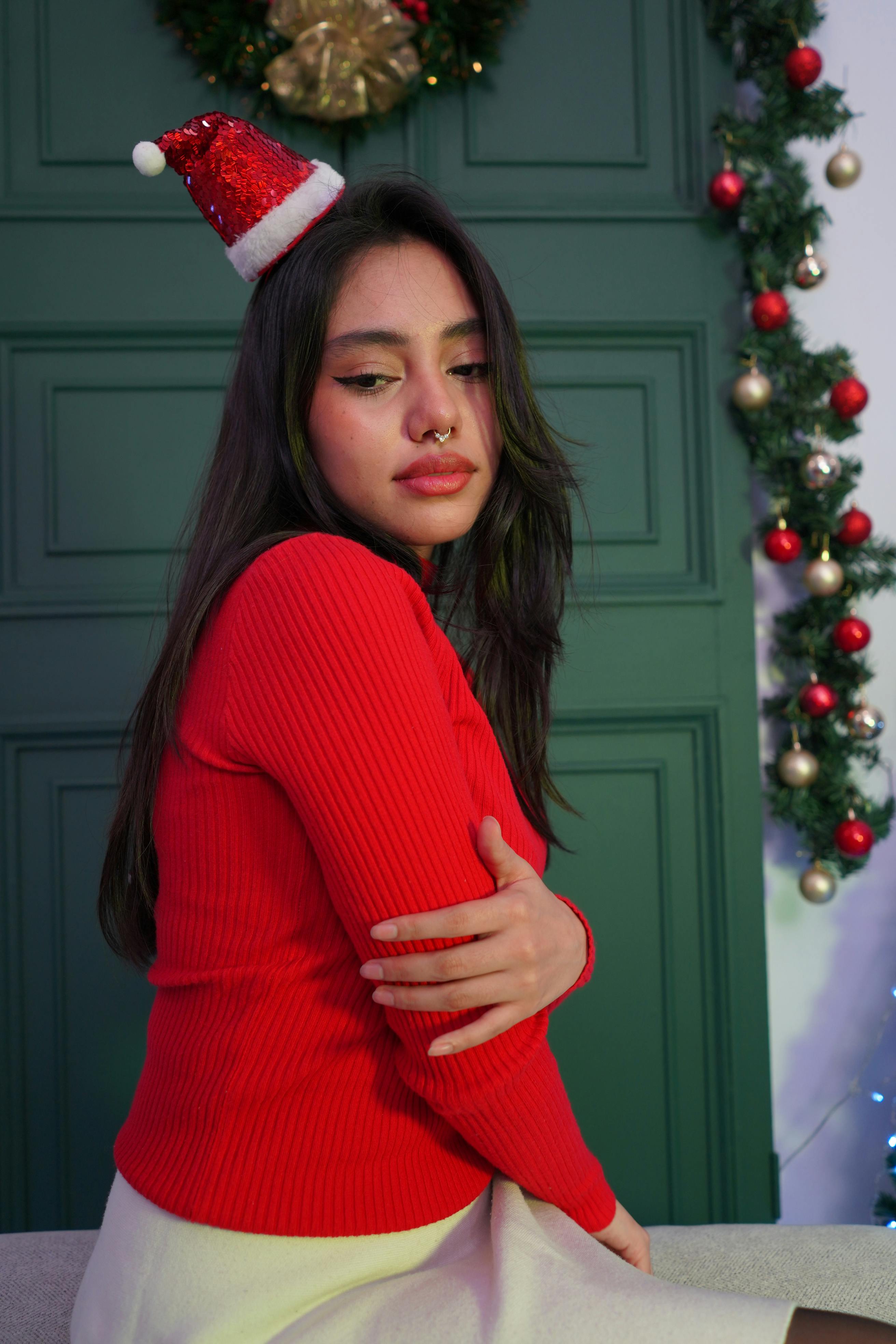 grátis Jovem mulher com suéter vermelho e gorro de Papai Noel posando em ambiente interno com decorações natalinas. Foto profissional