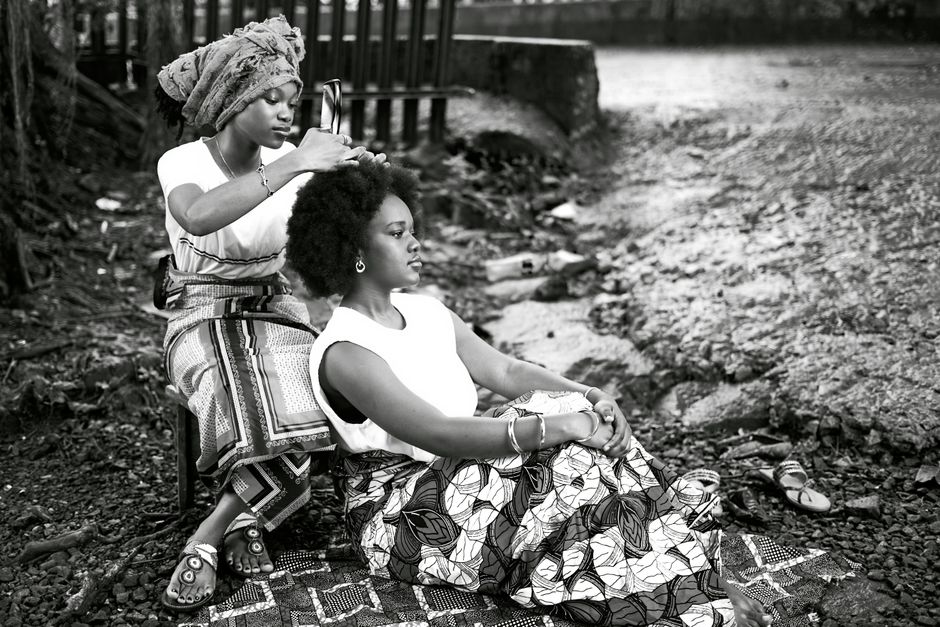 Two African women in traditional attire engage in hair styling outdoors, creating a cultural and intimate scene.