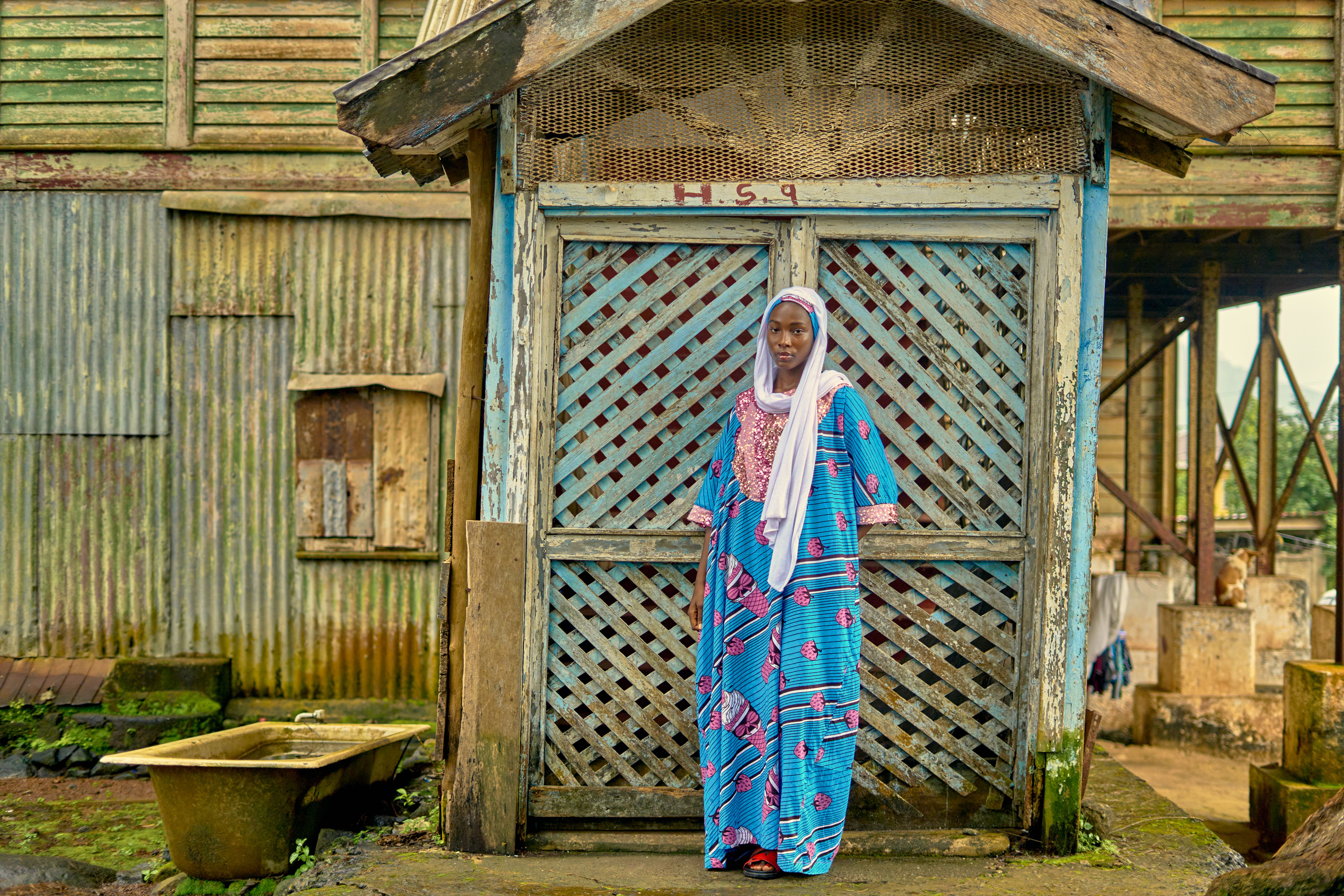 A woman in colorful traditional attire poses outdoors by a rustic wooden structure.
