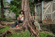 Two women sitting by a large tree in a lush, tropical environment with rustic architecture.