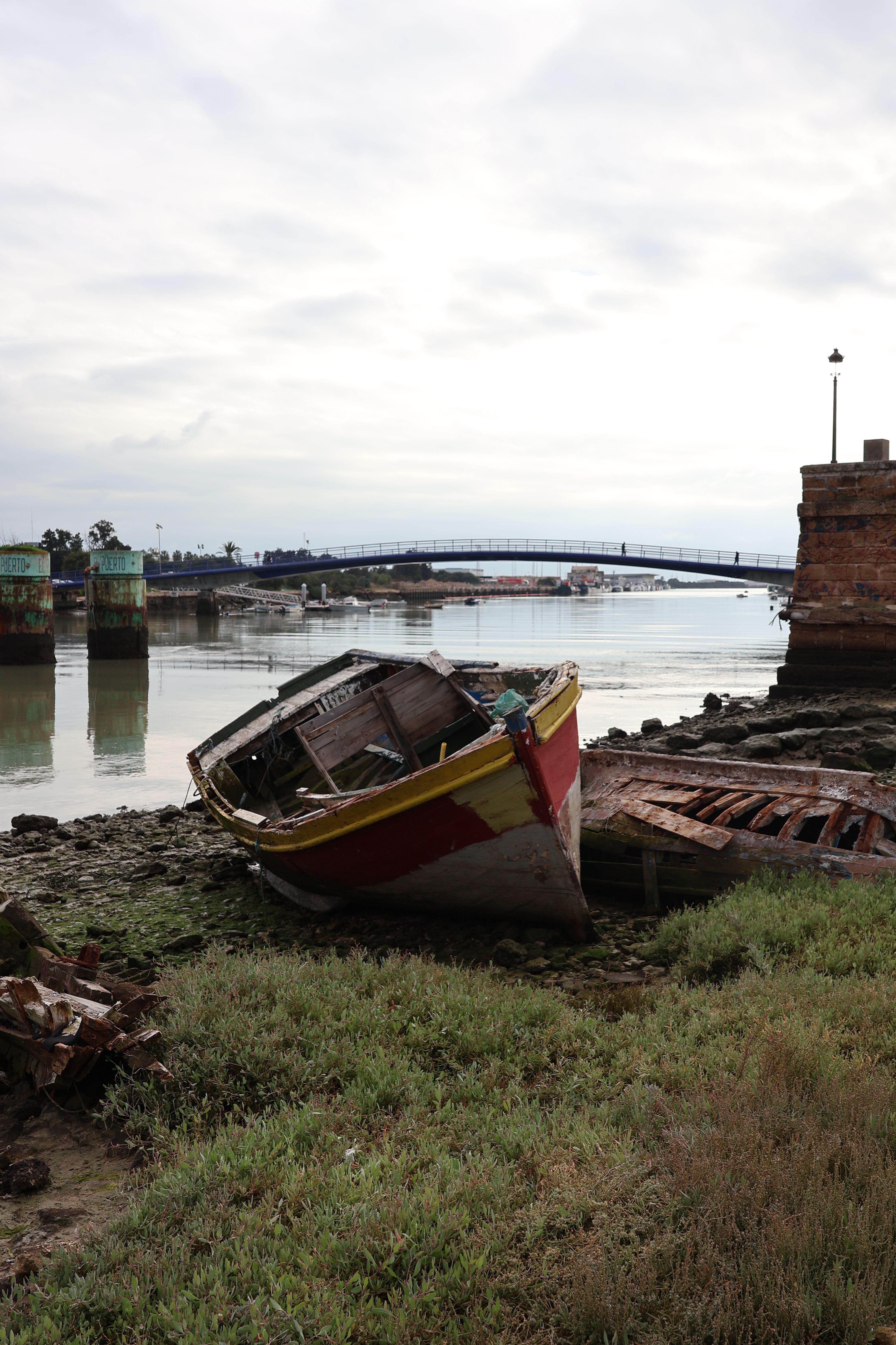 Weathered boat resting on a grassy riverbank with a distant bridge view.