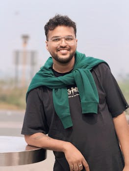 A smiling man in casual attire poses outdoors on a cloudy day, exuding a relaxed vibe.