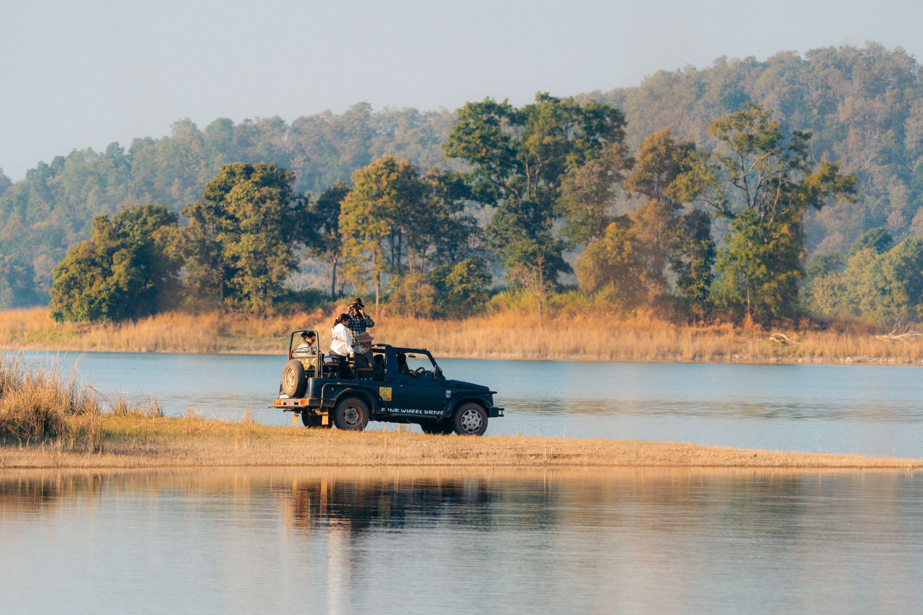 Explorers in a jeep enjoy a safari near a tranquil river surrounded by lush greenery.