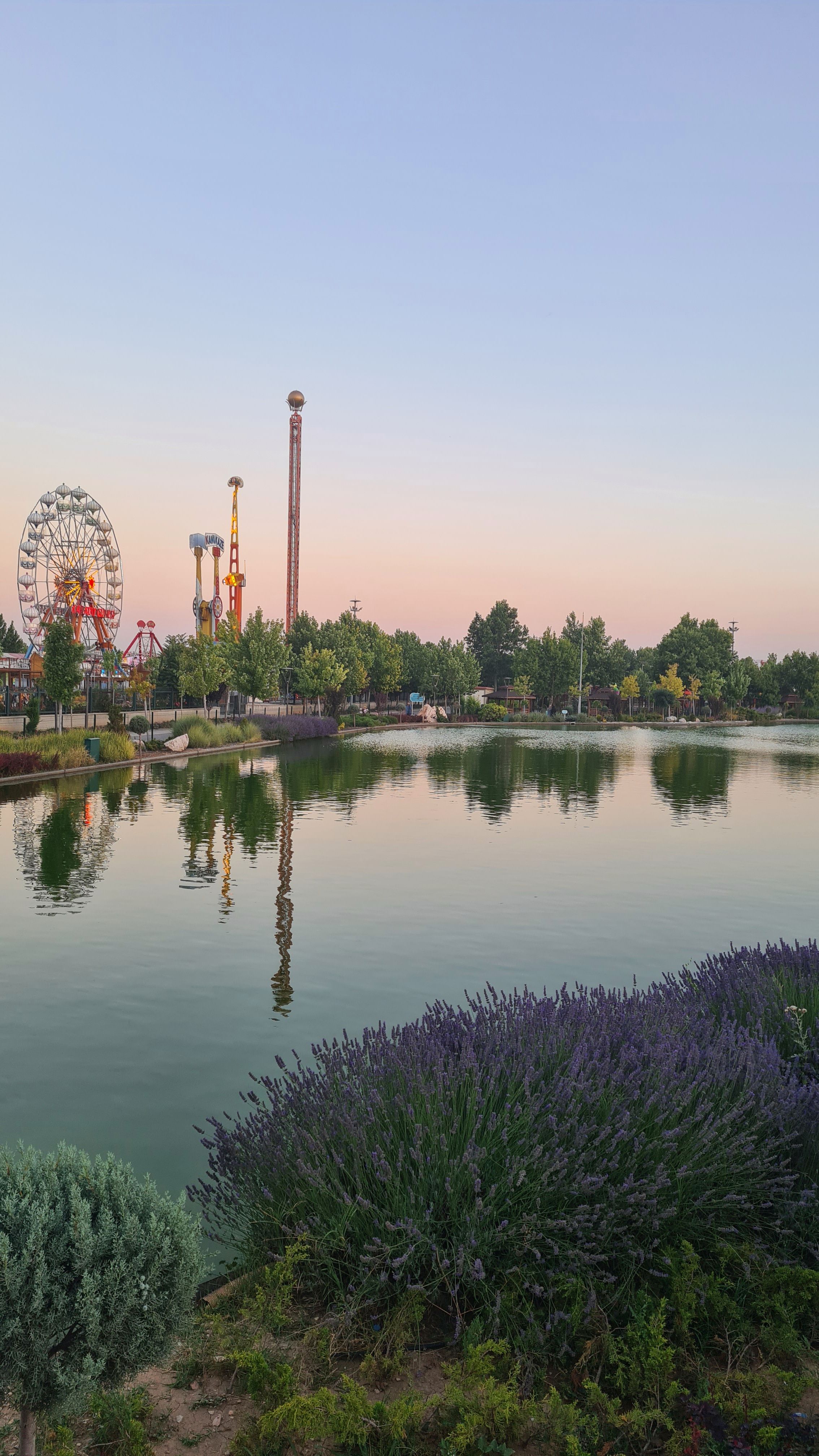 Calm lake reflections of an amusement park at sunset, featuring ferris wheel and rides.