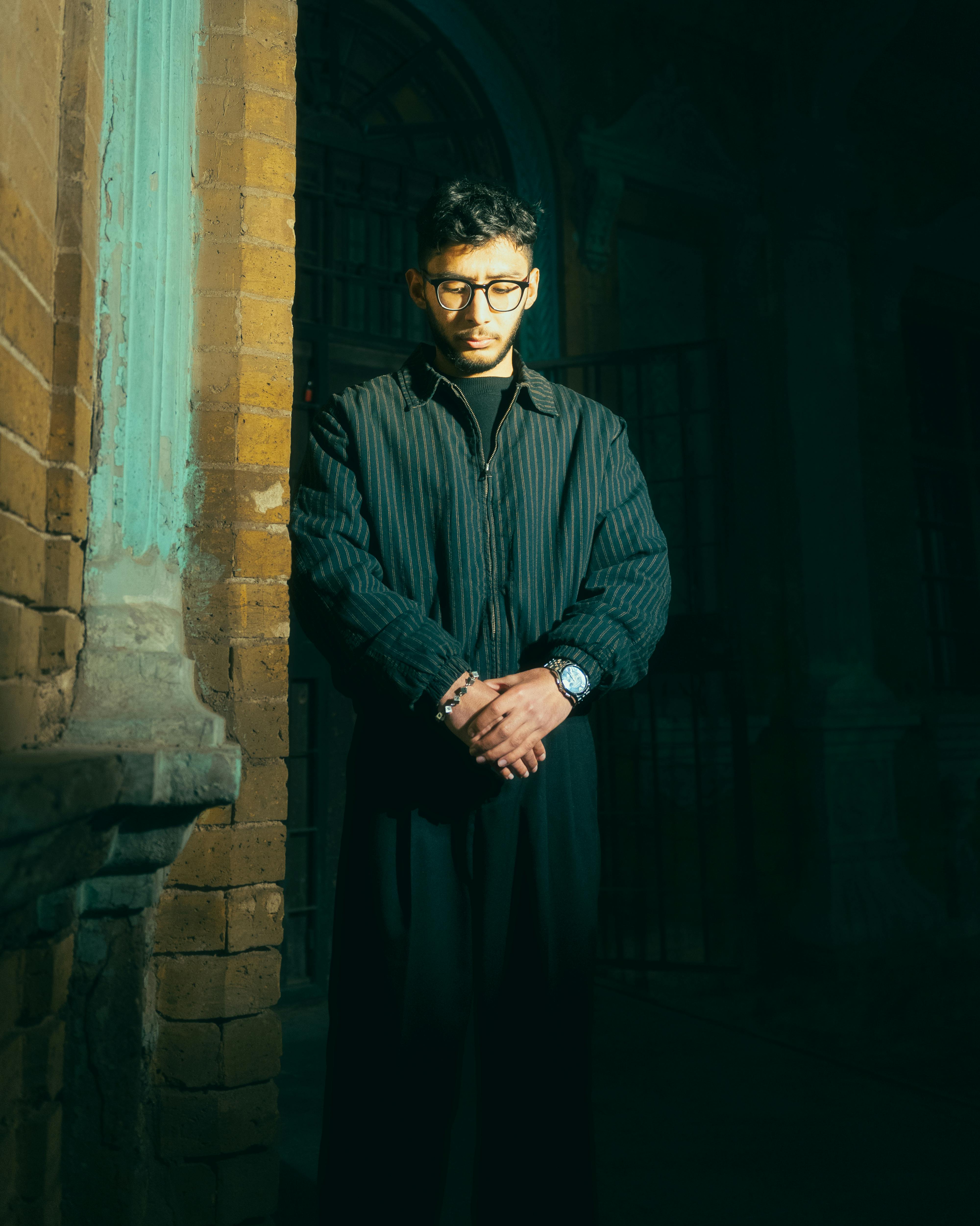 Gratis En mann med briller står høytidelig i et svakt opplyst miljø med stemningsfull, dramatisk belysning. Arkivbilde