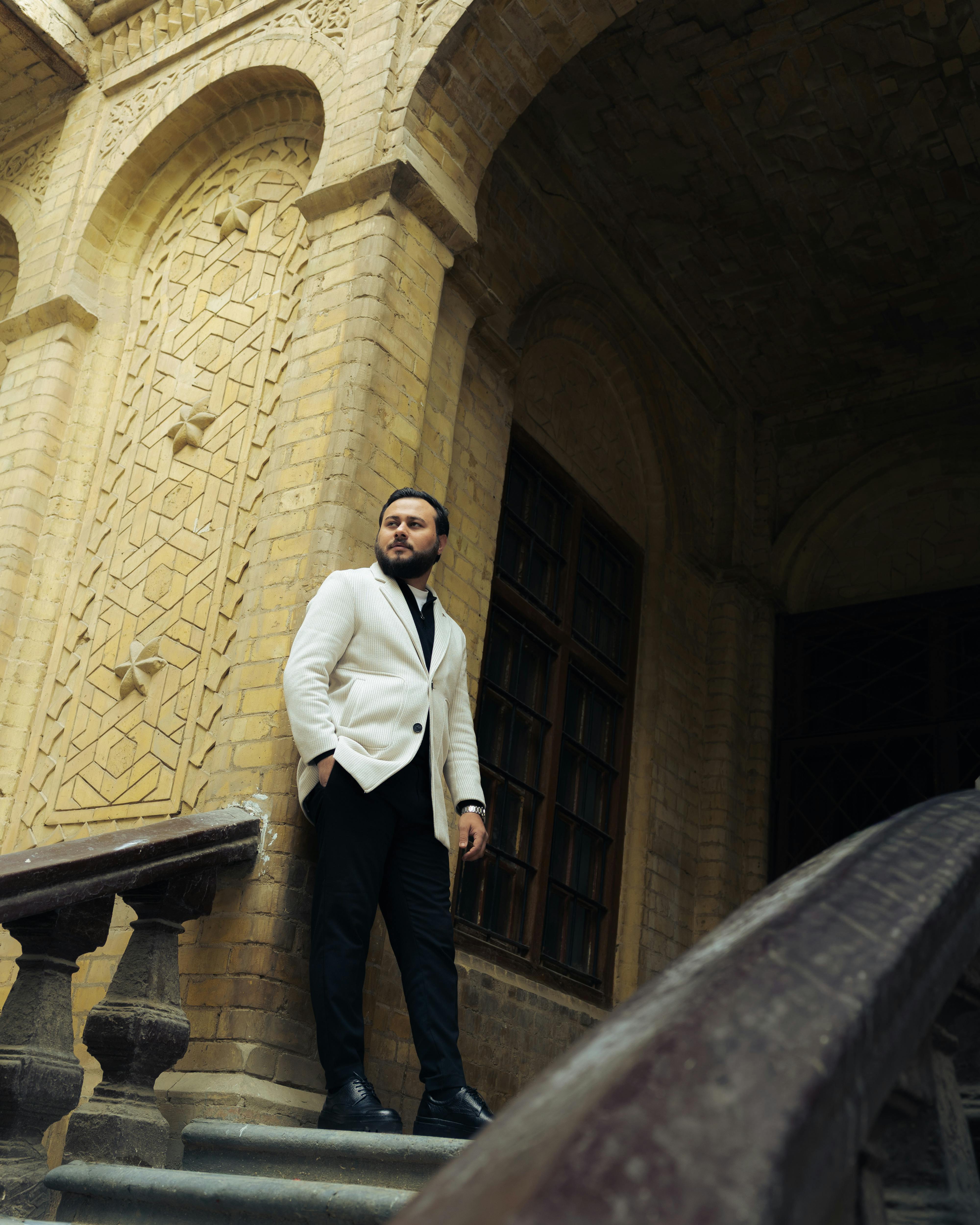 De franc Un home elegant amb jaqueta blanca es troba al costat d'un edifici de maó ornamentat, transmetent elegància. Foto d'estoc