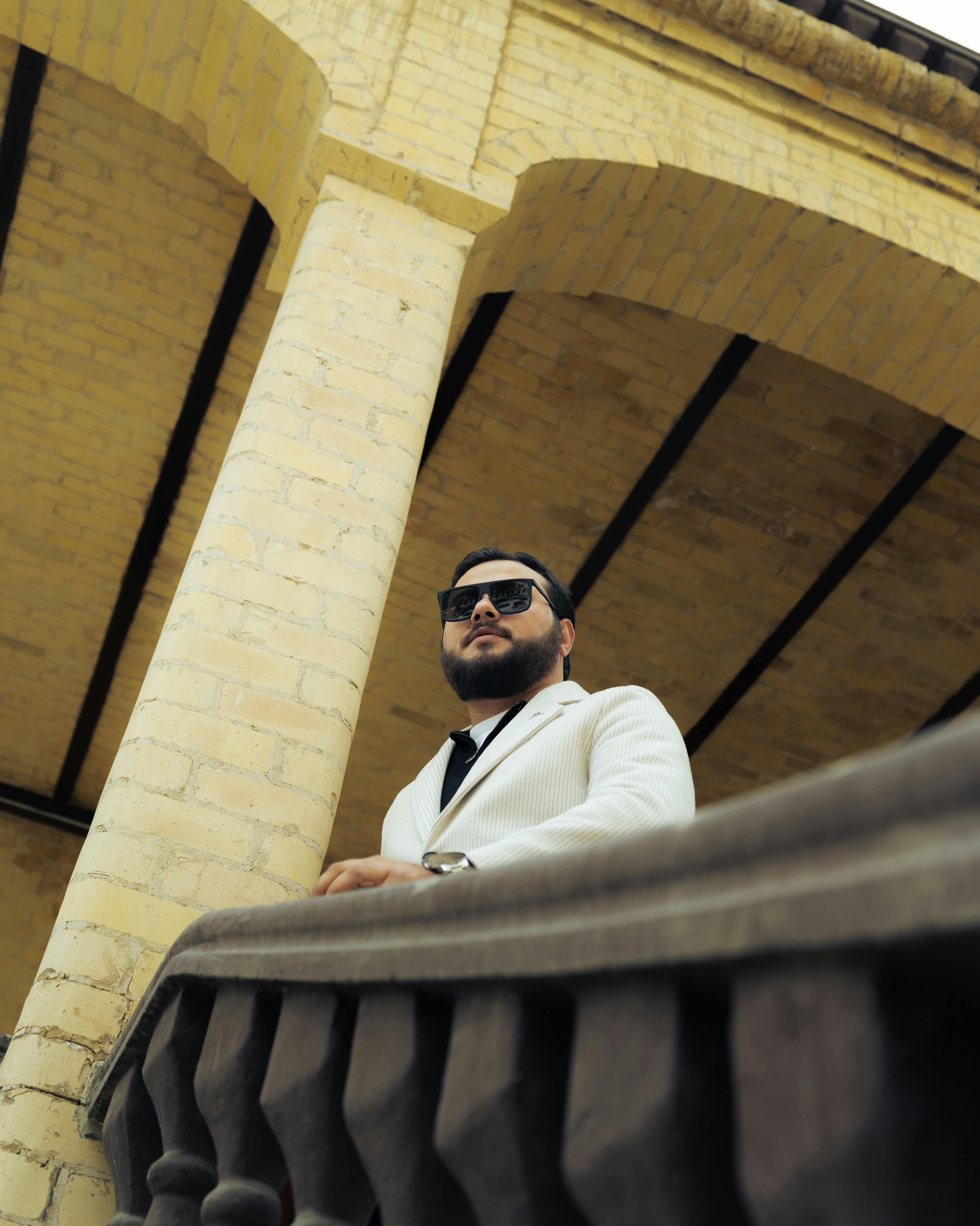 De franc Un home elegant amb ulleres de sol i vestit blanc es troba amb confiança en un balcó clàssic. Foto d'estoc