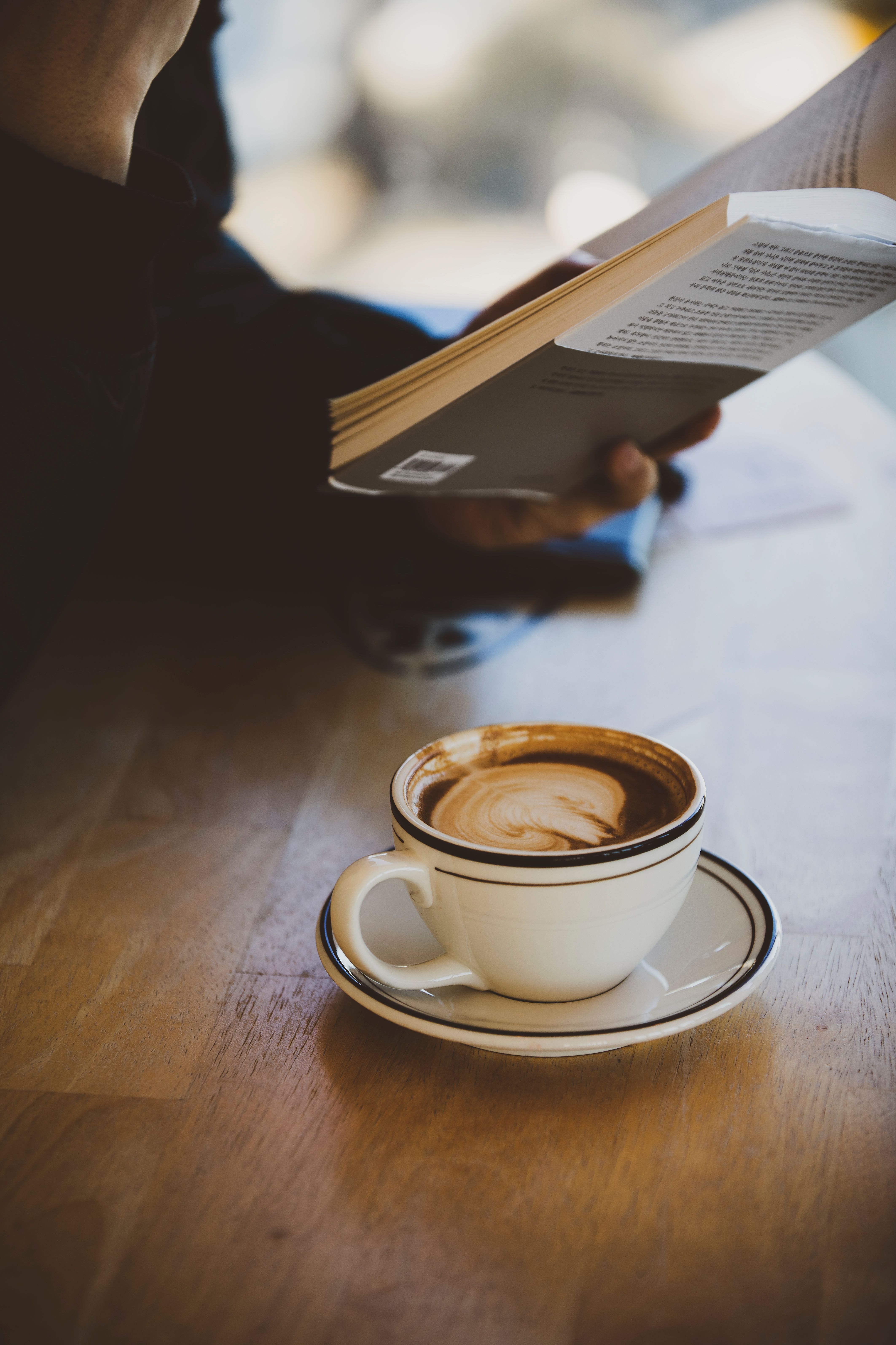 Latte art in cup on table with person reading a book, creating a peaceful atmosphere.