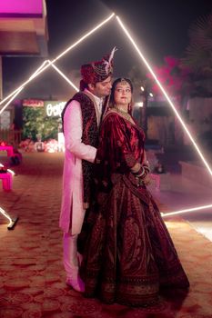 A couple in traditional Indian wedding attire poses under geometric lighting.