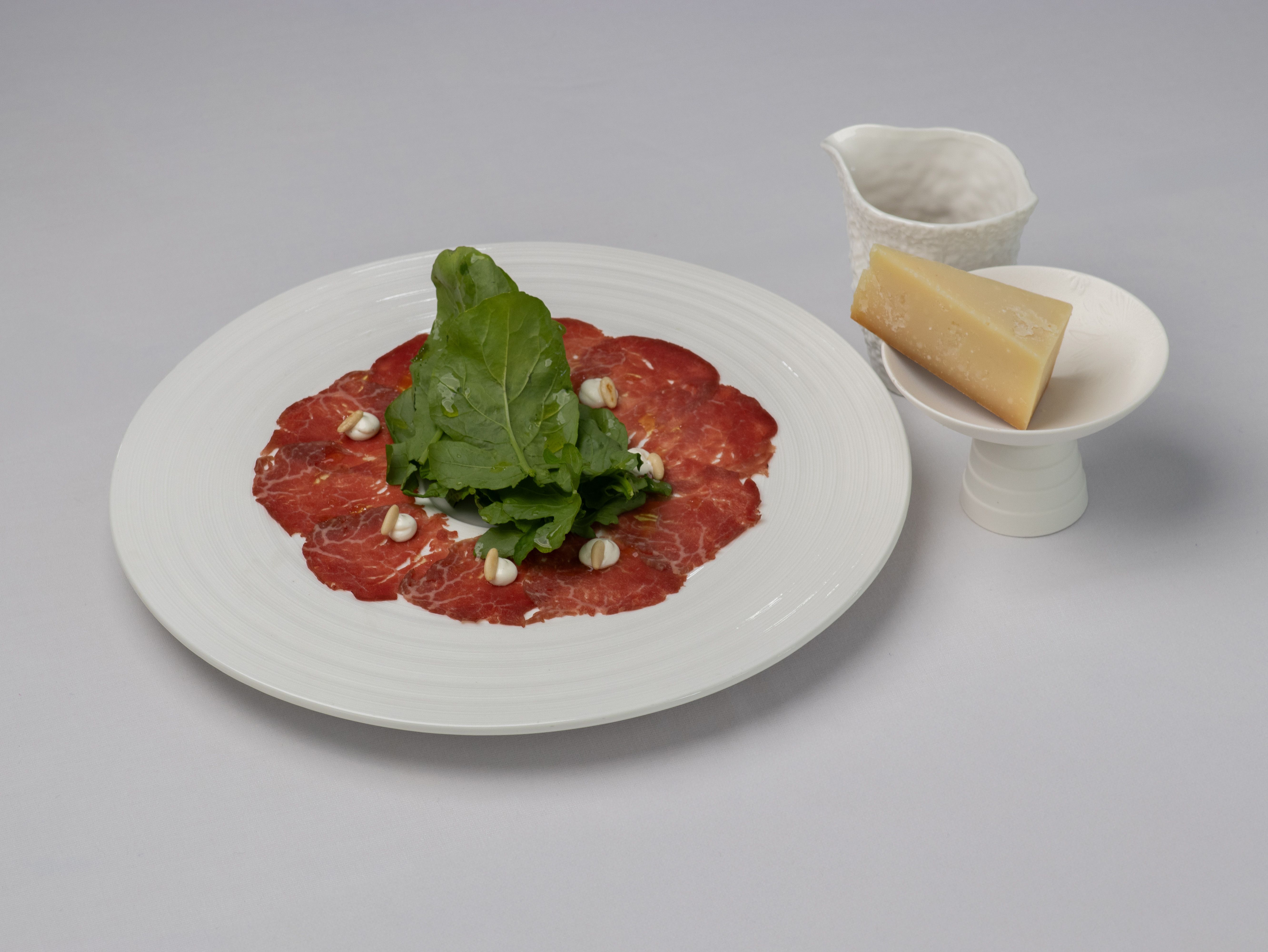 Formal beef carpaccio plated with arugula, Parmesan, and a drizzle jug, offering a sophist