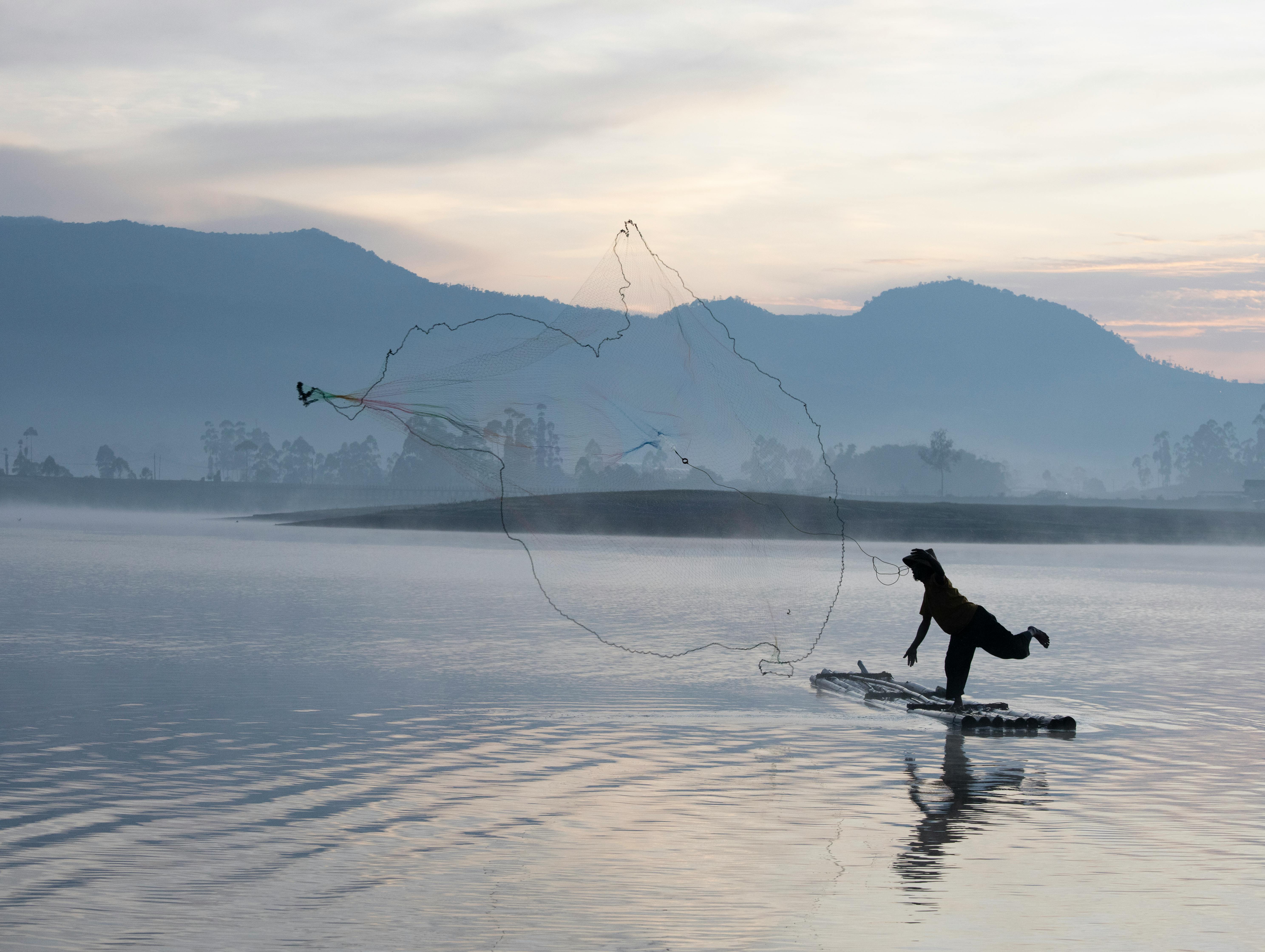Gratuit Silhouette d'un pêcheur jetant ses filets au lever du soleil sur un lac serein à Java, en Indonésie. Photos