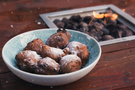 Delicious Dutch oliebollen dusted with powdered sugar in a bowl by a fire pit.