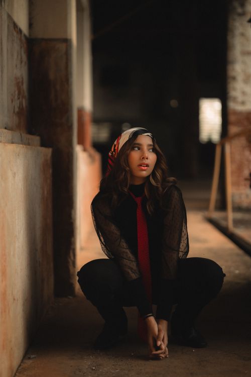 Free Fashionable young woman crouching with headscarf in moody indoor setting. Stock Photo