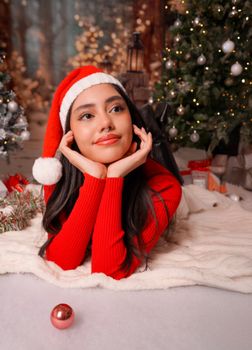 Young woman in Santa hat lying by a Christmas tree, surrounded by festive decor.