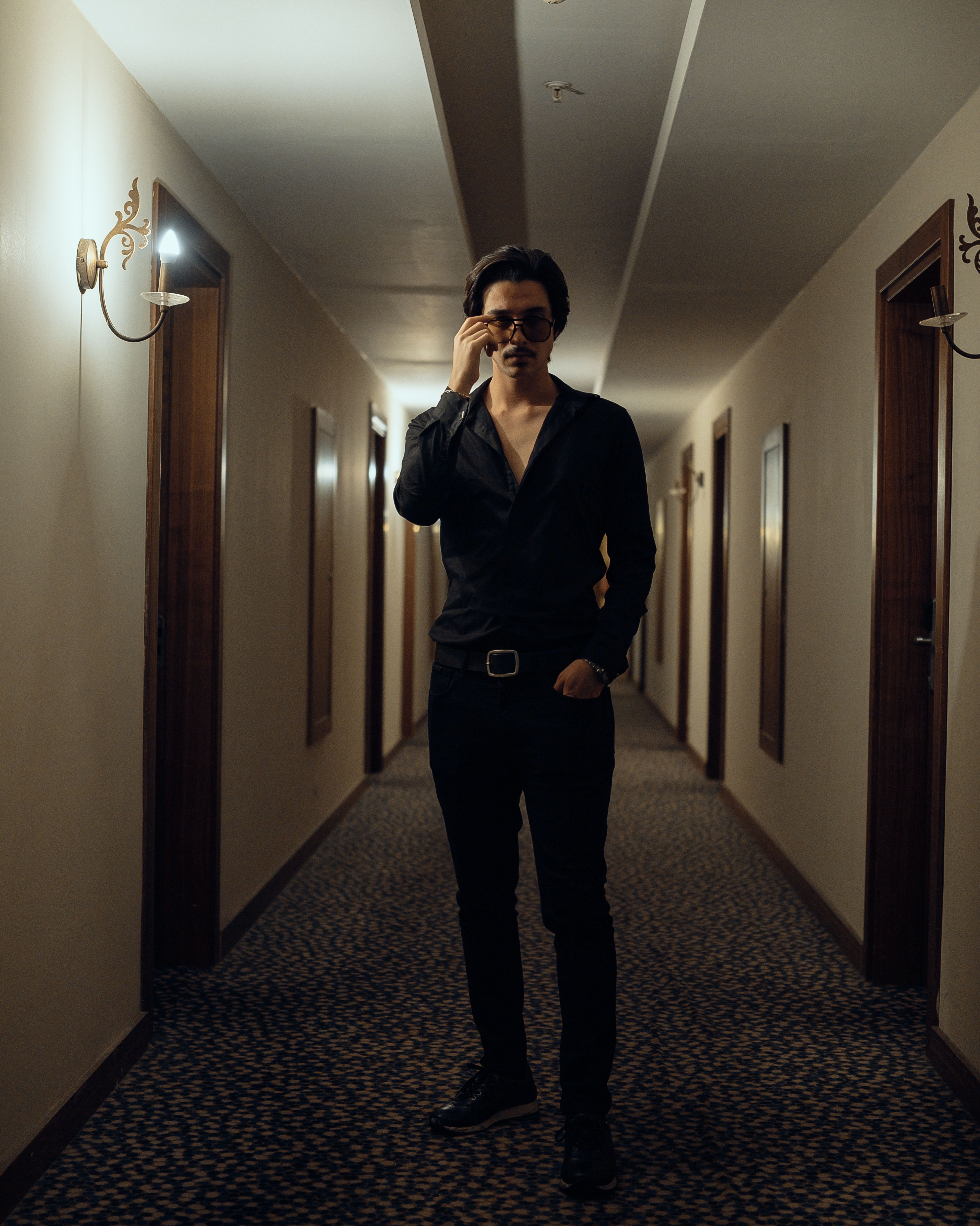Confident man in black outfit walking through a dimly lit hotel corridor, stylish and poised.