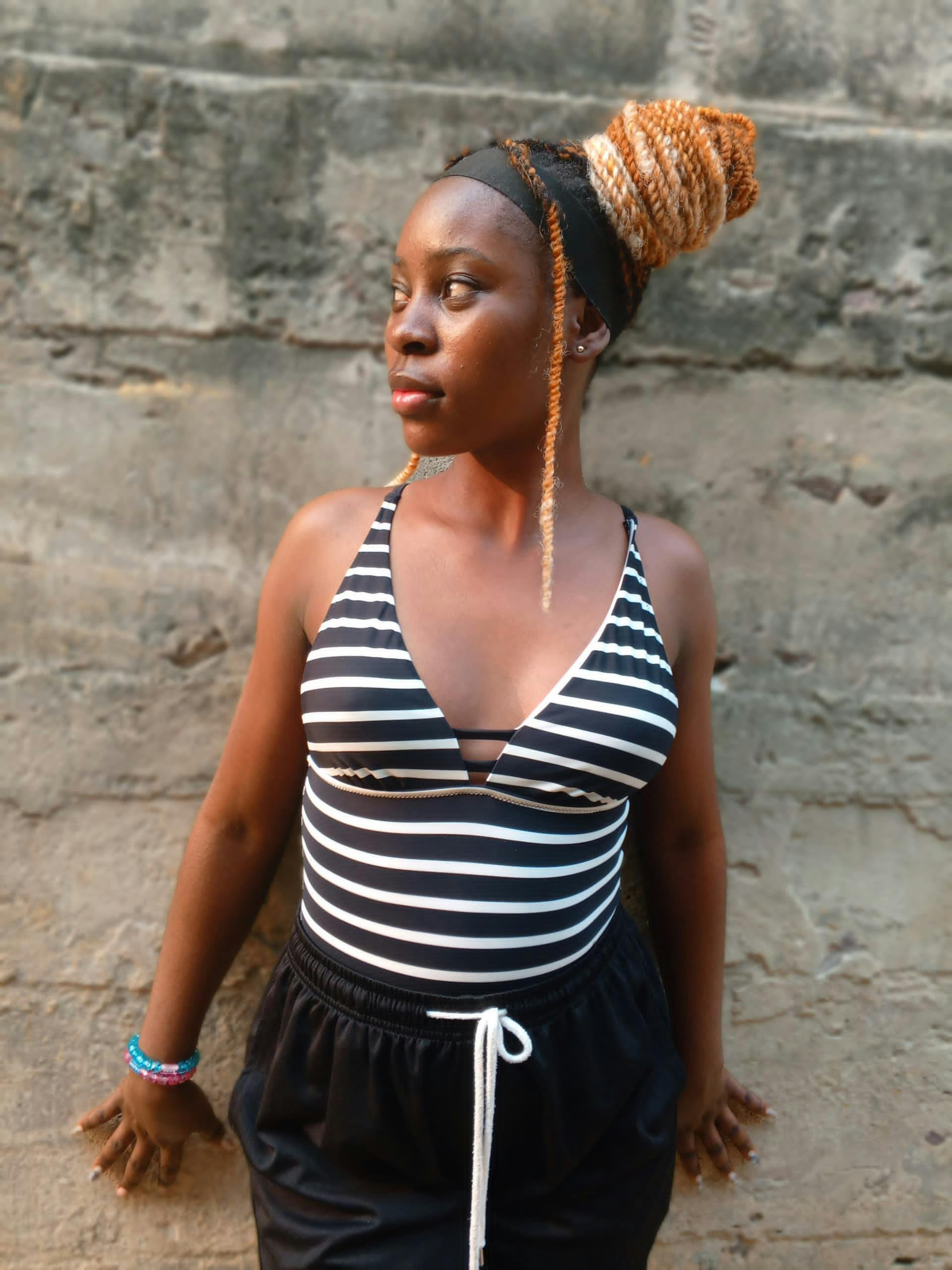 Gratis Una mujer joven en traje de baño a rayas mira con confianza hacia una pared rústica, exudando fuerza y estilo. Foto de stock