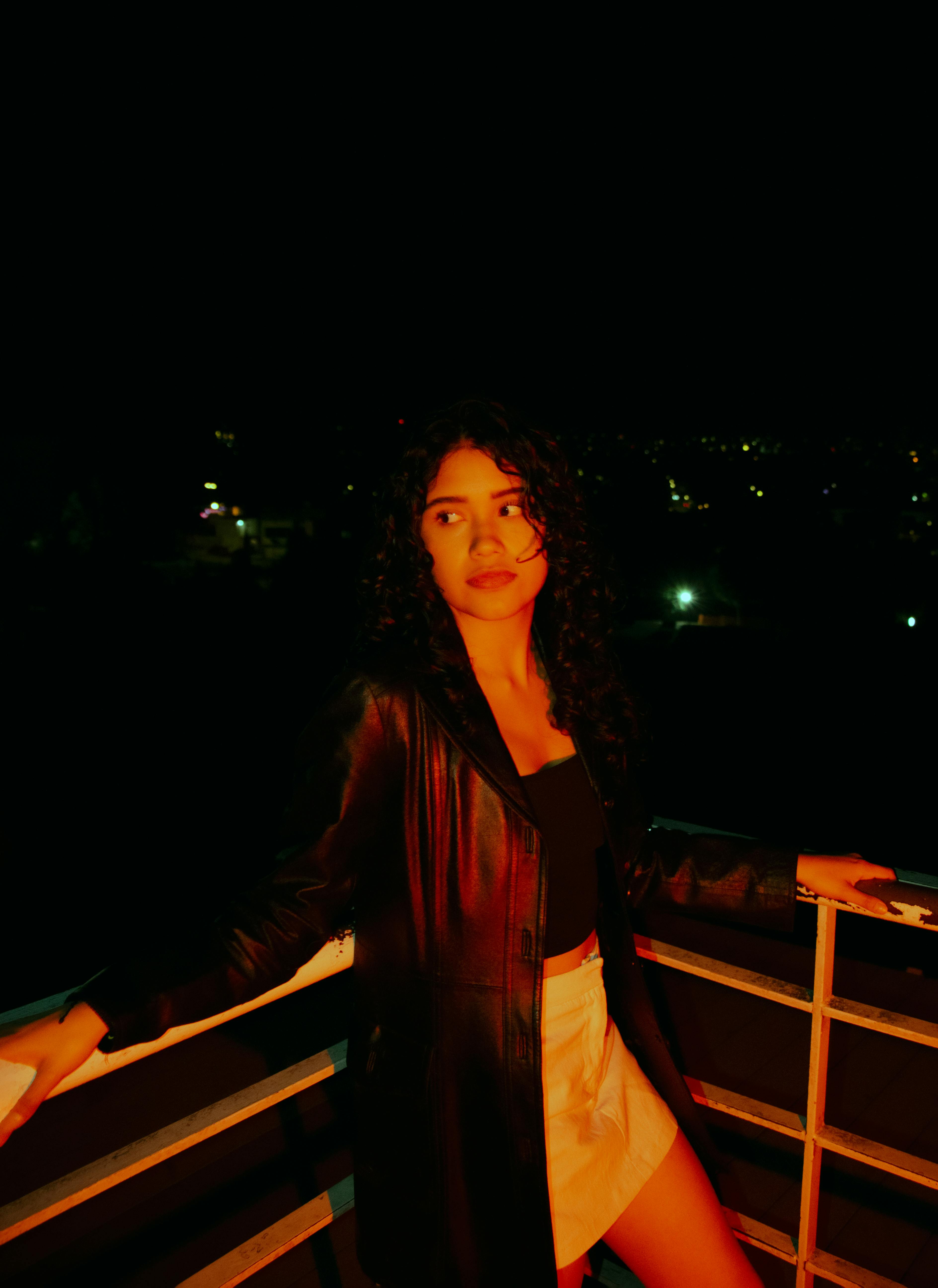 De franc Una dona jove amb jaqueta de cuir està dreta en una teulada, amb els llums de la ciutat al fons. Foto d'estoc