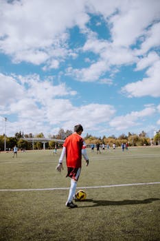 A teenage soccer player in red preps to kick a ball during a match on a sunny field.