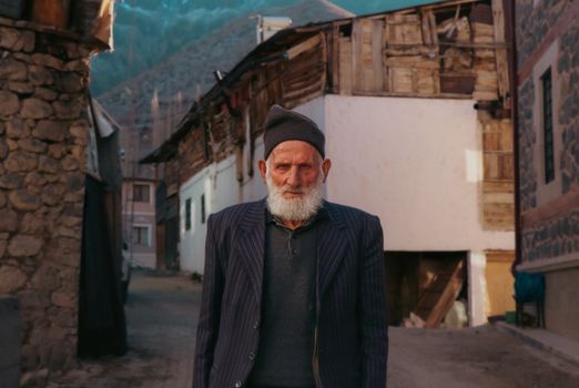 Bearded elderly man in a village street with rustic architecture and mountainous background.