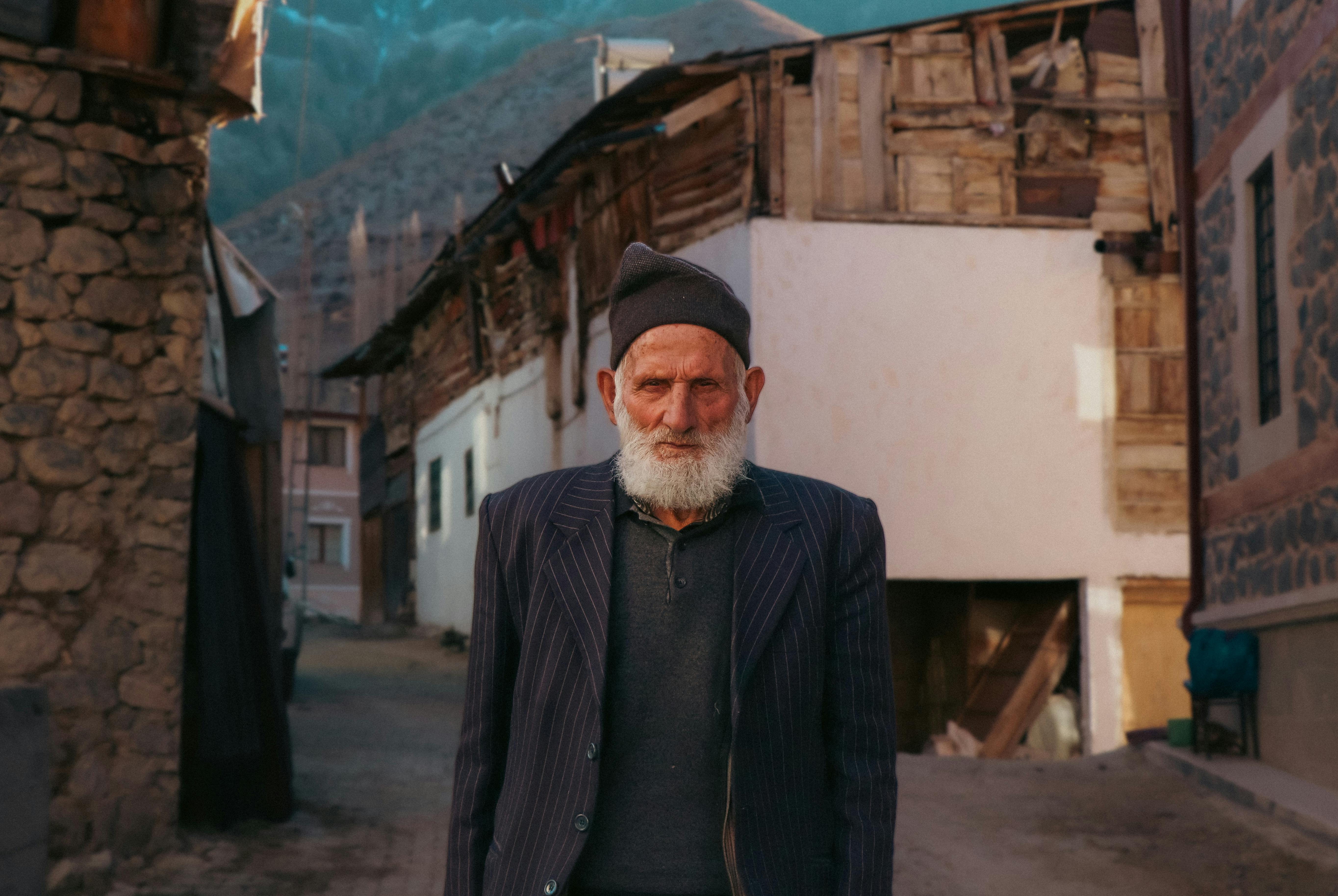 Gratis Skjeggete eldre mann i en landsbygate med rustikk arkitektur og fjellrik bakgrunn. Arkivbilde