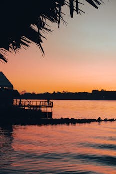 A picturesque lakeside view at sunset featuring silhouetted structures and vibrant colors.