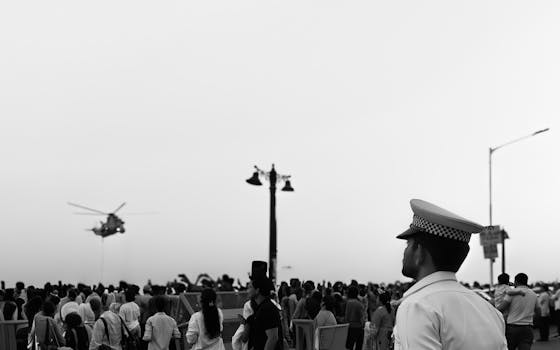Crowd gathered outdoors watching a hovering helicopter with a police officer in foreground.