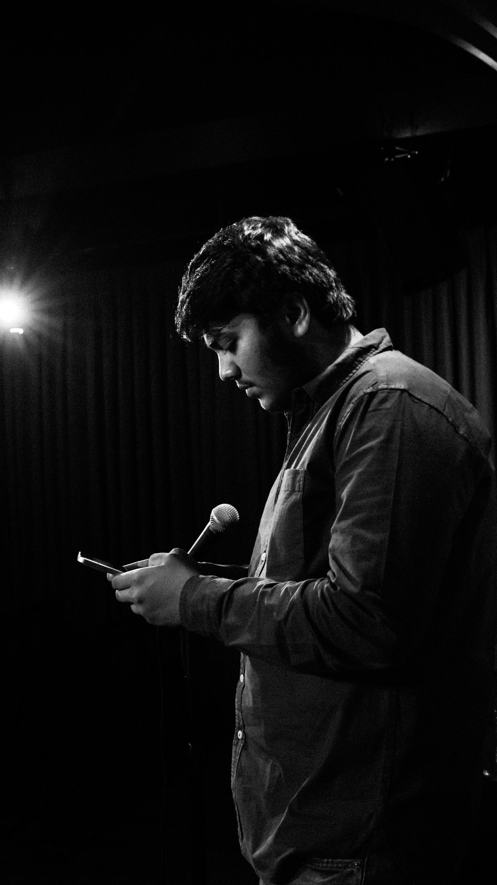 A comedian holding a microphone on stage, captured in a dramatic black and white setting.