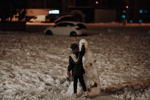 Free Young child building a snowman in snowy urban area at night with streetlights Stock Photo