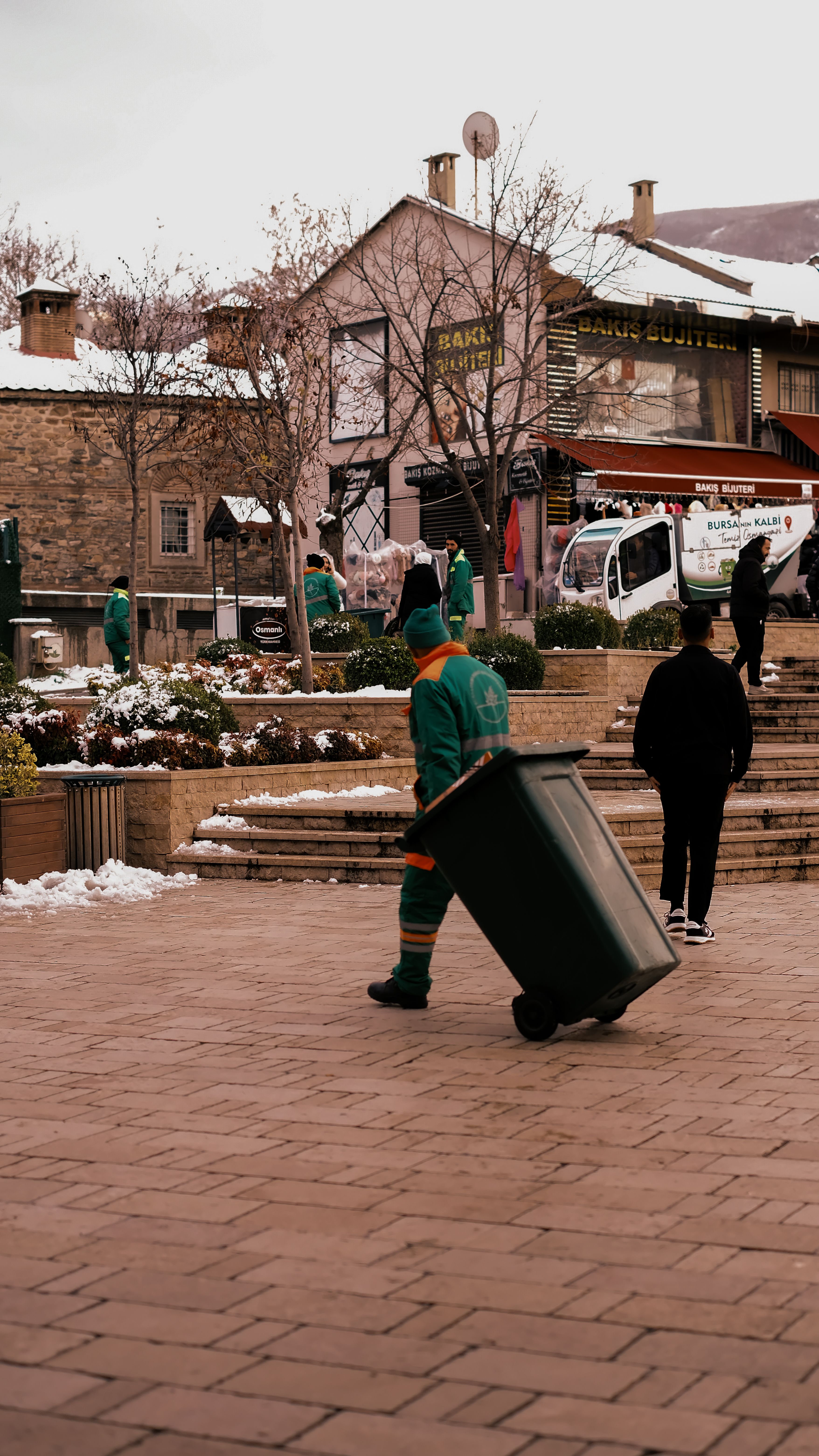 Sanitation workers in winter attire cleaning a cobblestone street lined with buildings.