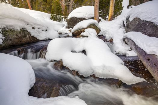A serene winter scene of a stream flowing between snow-covered rocks and trees in a forest.