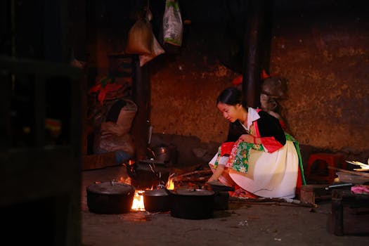 A woman in colorful attire cooks indoors over an open flame, highlighting traditional methods.