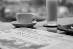 Elegant black and white photo of a cup of coffee, juice, and newspaper on a table in a serene setting.