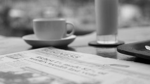 Elegant black and white photo of a cup of coffee, juice, and newspaper on a table in a serene setting.