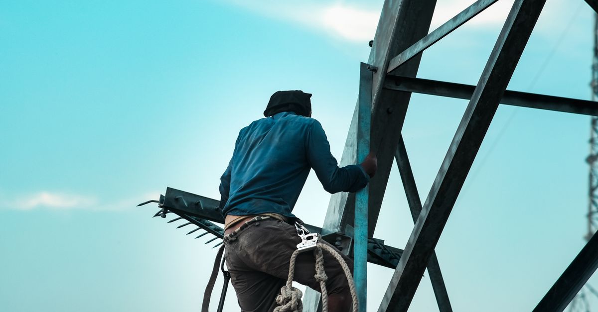 A construction worker climbs a steel tower with safety gear against a clear sky in Mymensingh, Bangladesh.