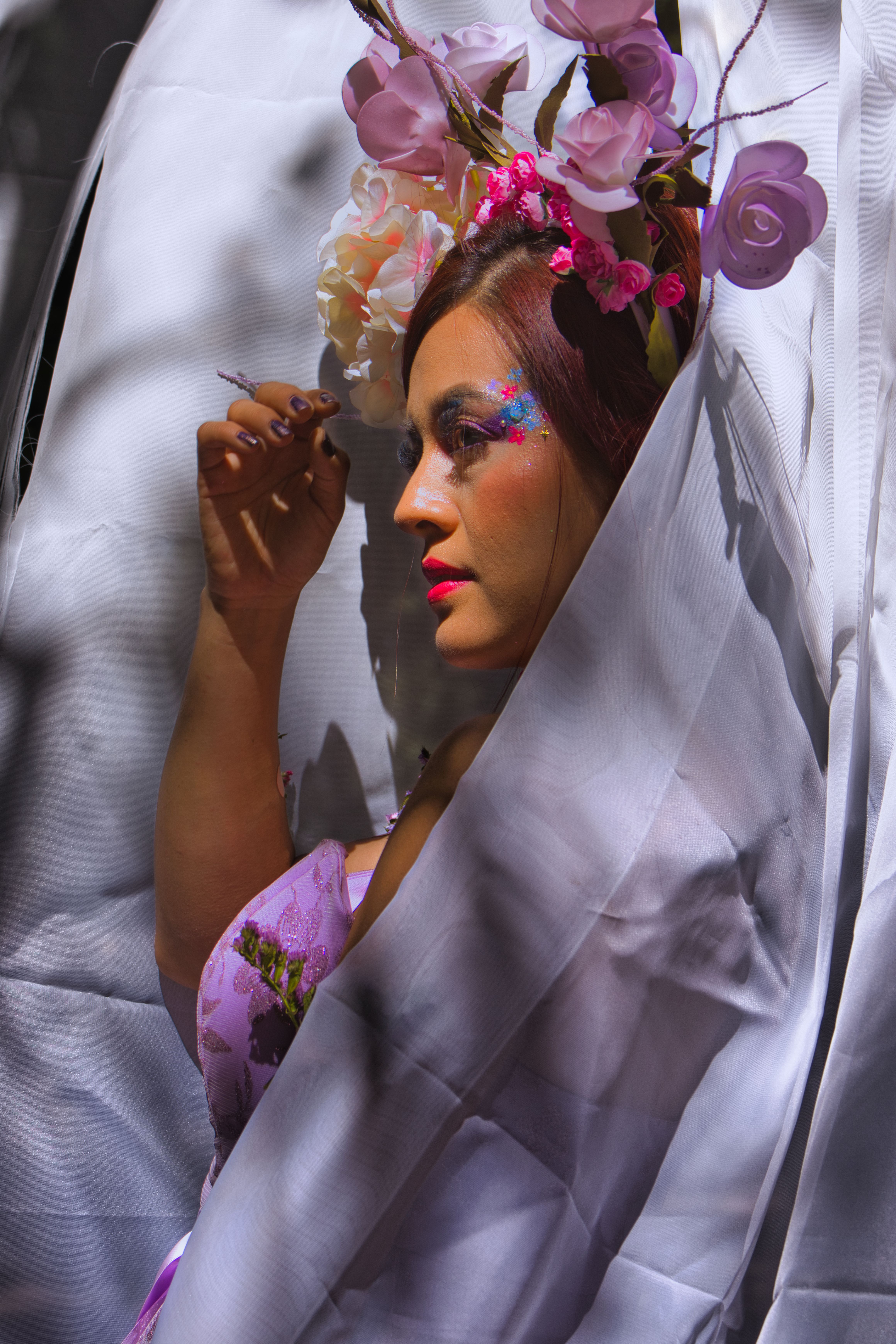 Portrait of a woman in floral headpiece with colorful glitter makeup through soft white fabric.