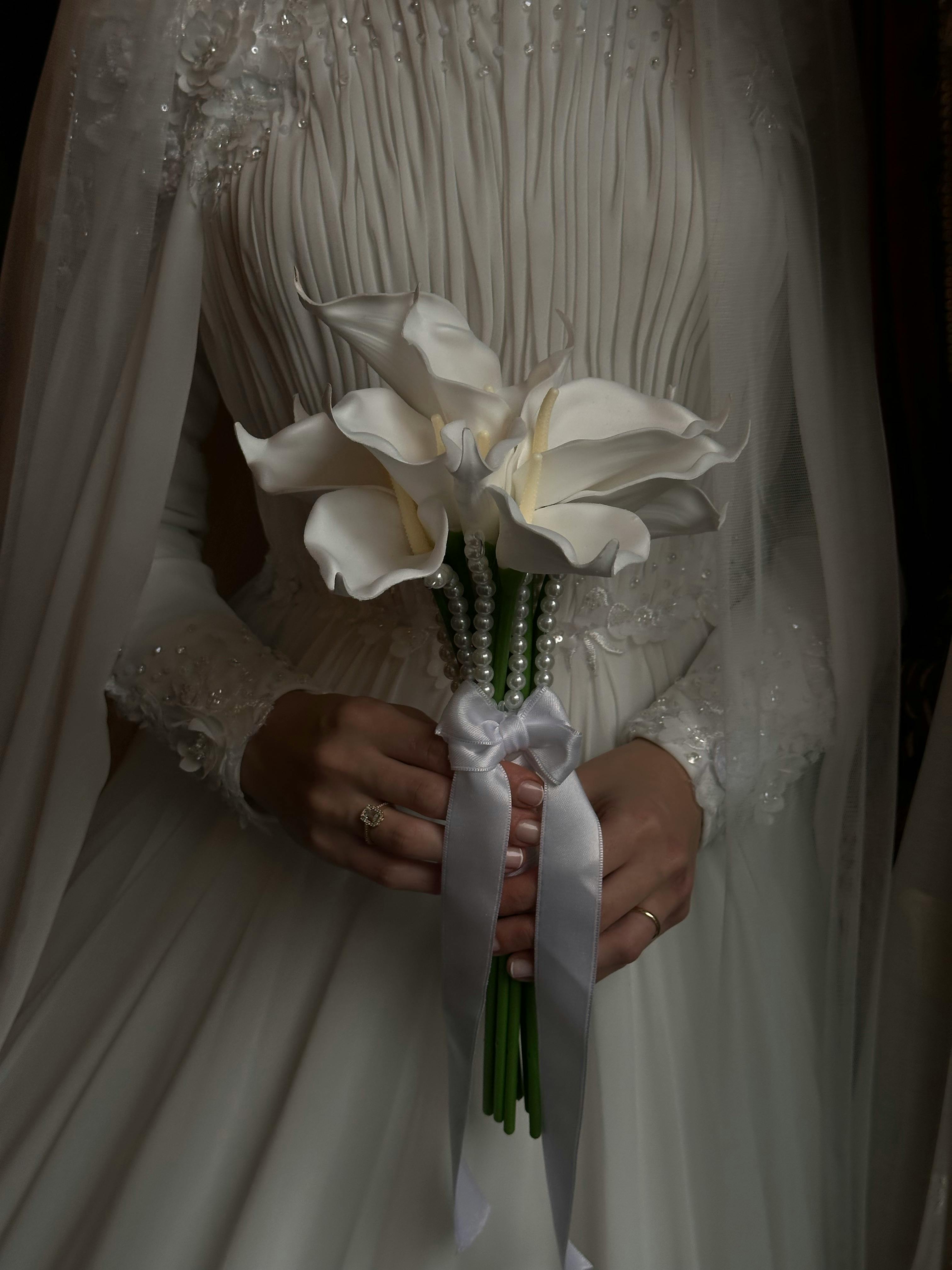 Kostenlos Braut in weißem Kleid, die einen mit Perlen und Bändern verzierten Calla-Lilien-Strauß hält. Stock-Foto