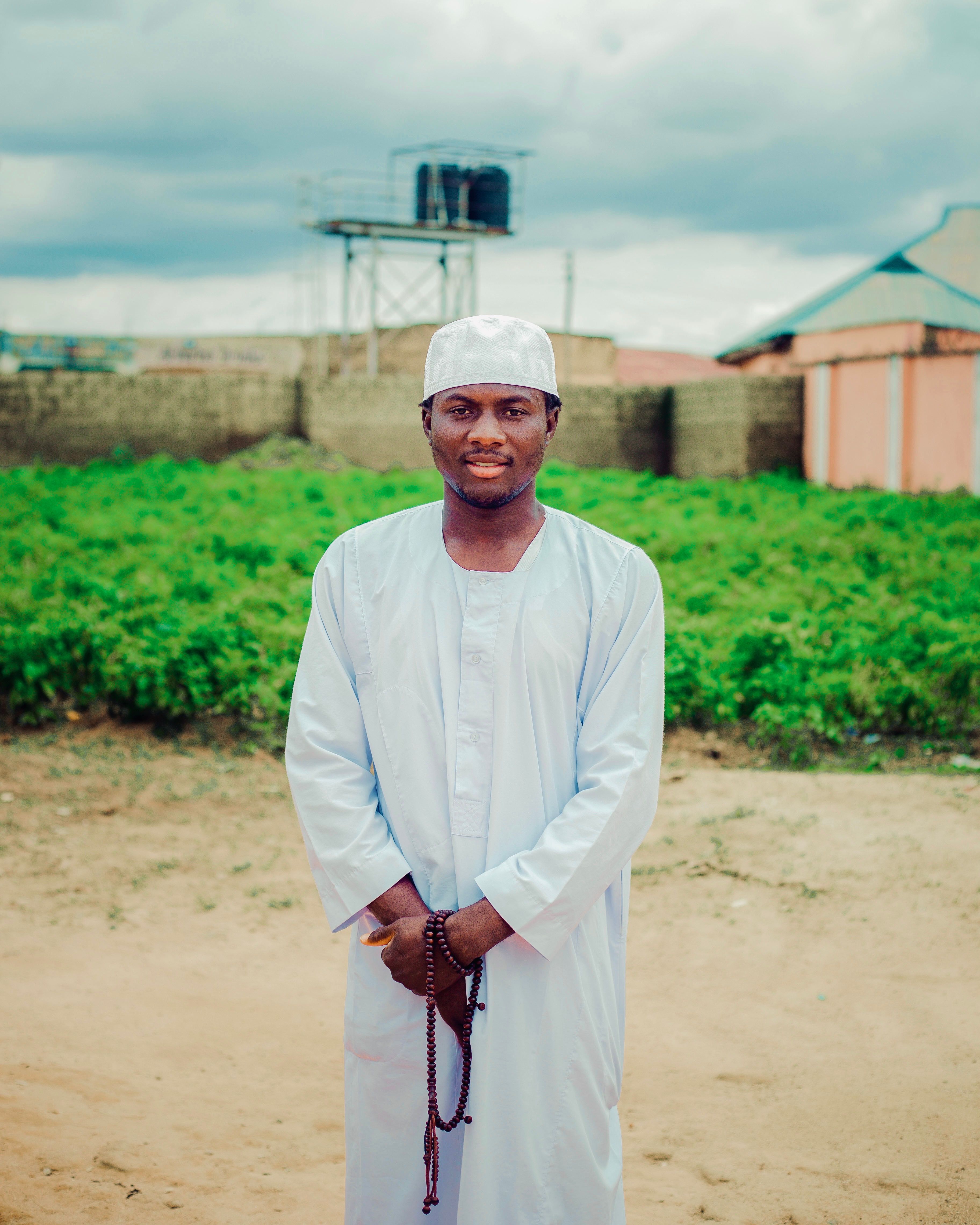 African man in traditional white attire outdoors by a green field, serene expression.