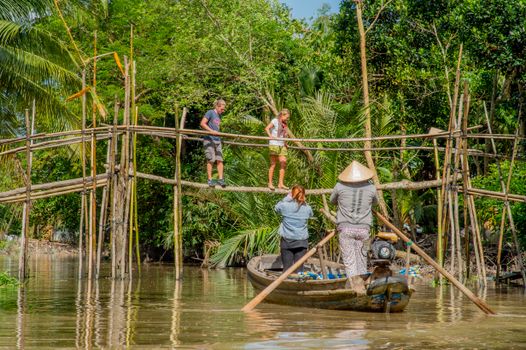 Tourists experience traditional bamboo bridge crossing in a lush tropical area with a local guide in a boat.