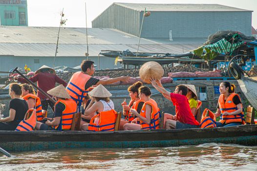 Tourists wearing life jackets and conical hats explore a vibrant floating market in a traditional boat.
