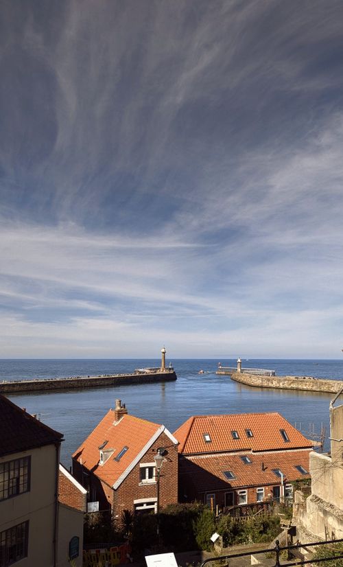 Free Beautiful view of Whitby Harbor with iconic red rooftops and lighthouse under a dramatic sky. Stock Photo
