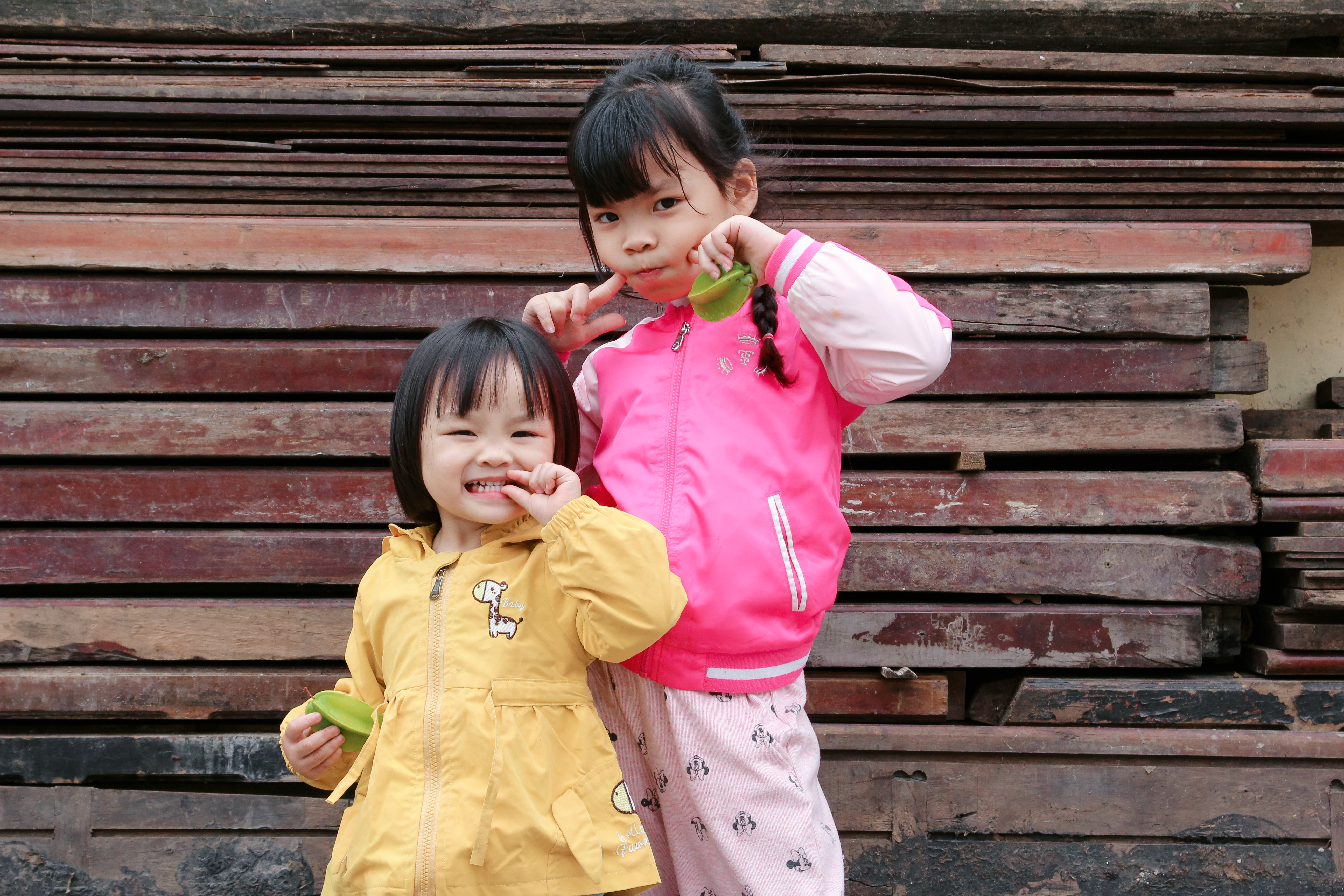 Two cheerful children playing with leaves in front of a wooden backdrop, showcasing joy and friendship.