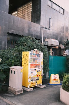 Yellow vending machine with Pokémon design on city sidewalk, surrounded by greenery.