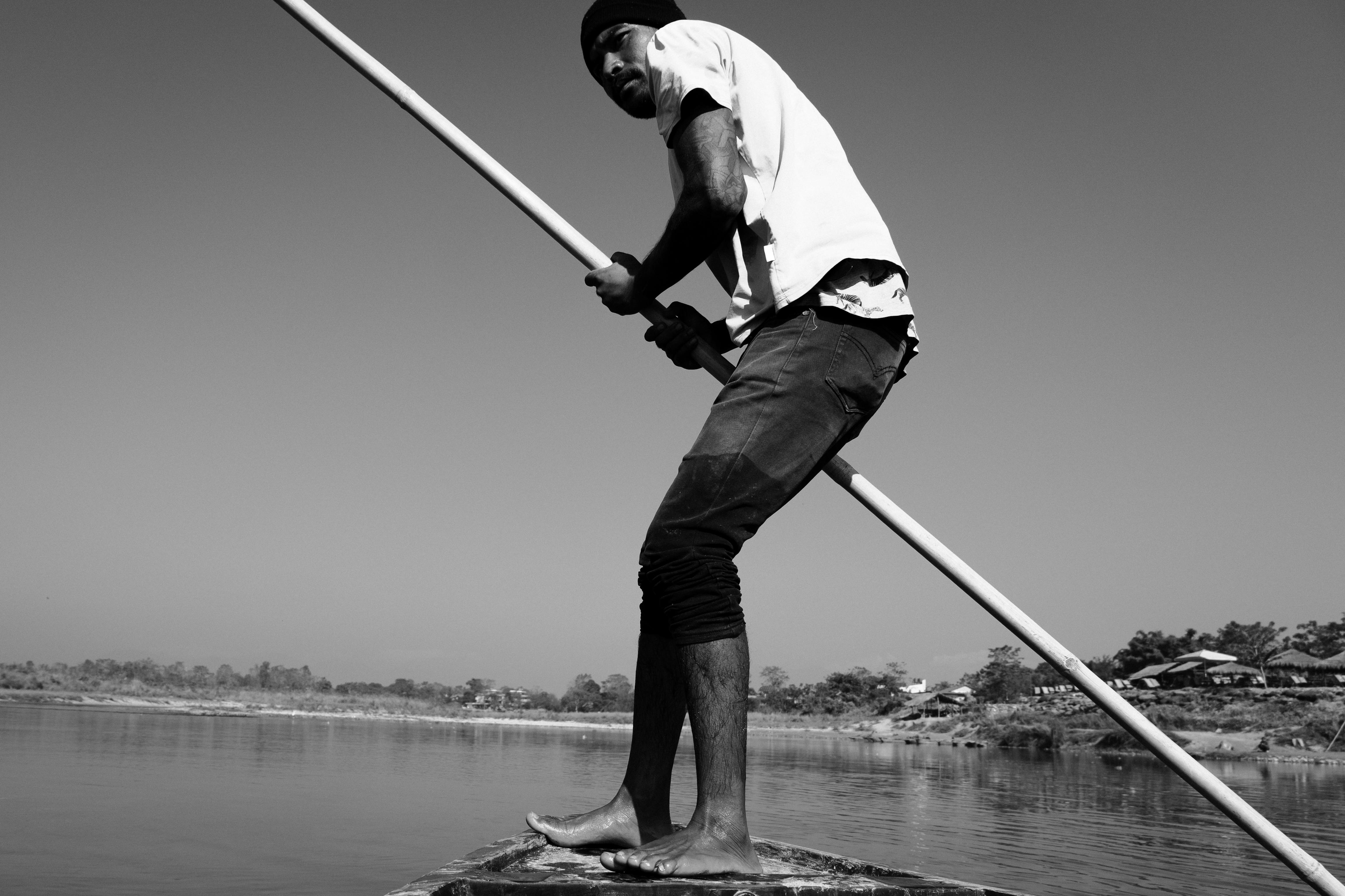 Black and white image of a man paddling a boat on a serene river under clear skies.
