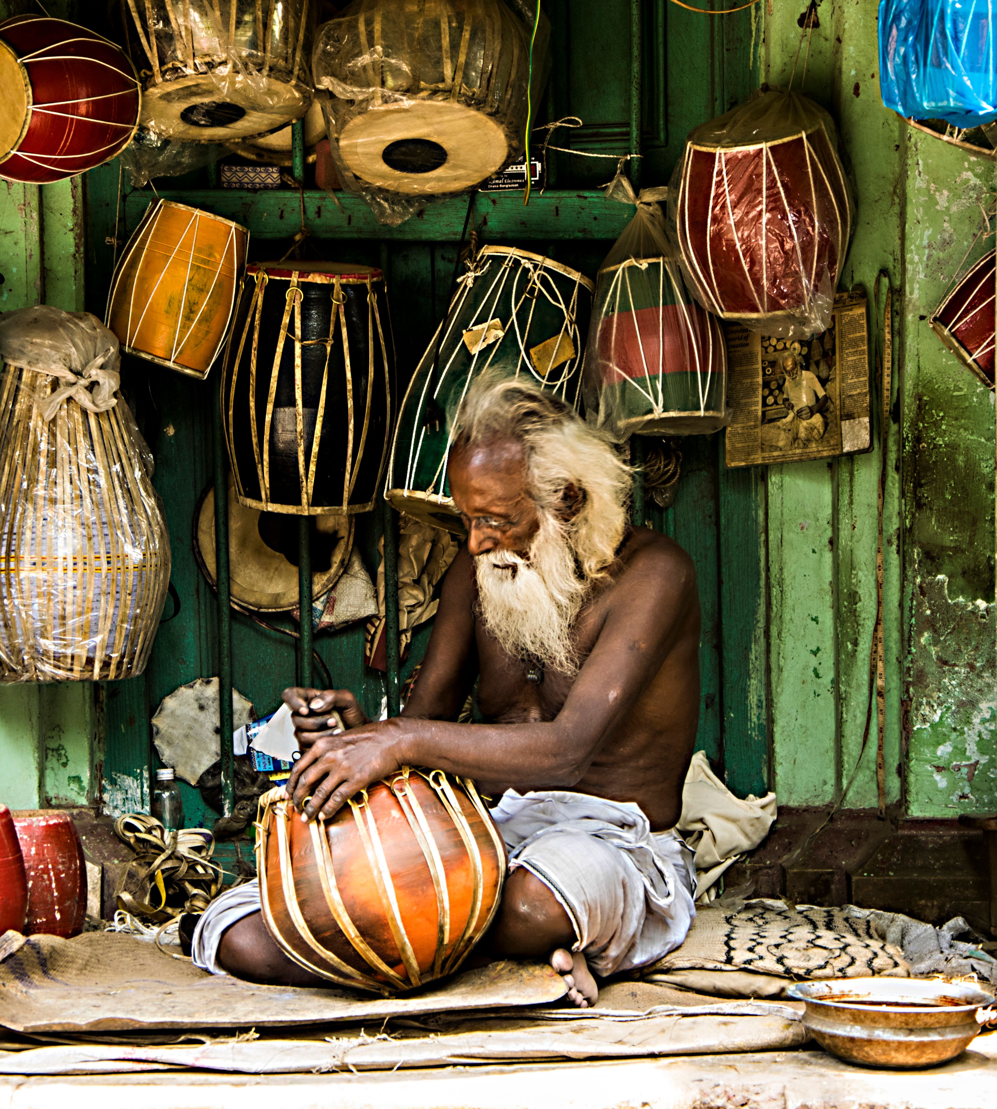 An elderly artisan working on crafting traditional Indian drums in his workshop filled with percussion instruments.