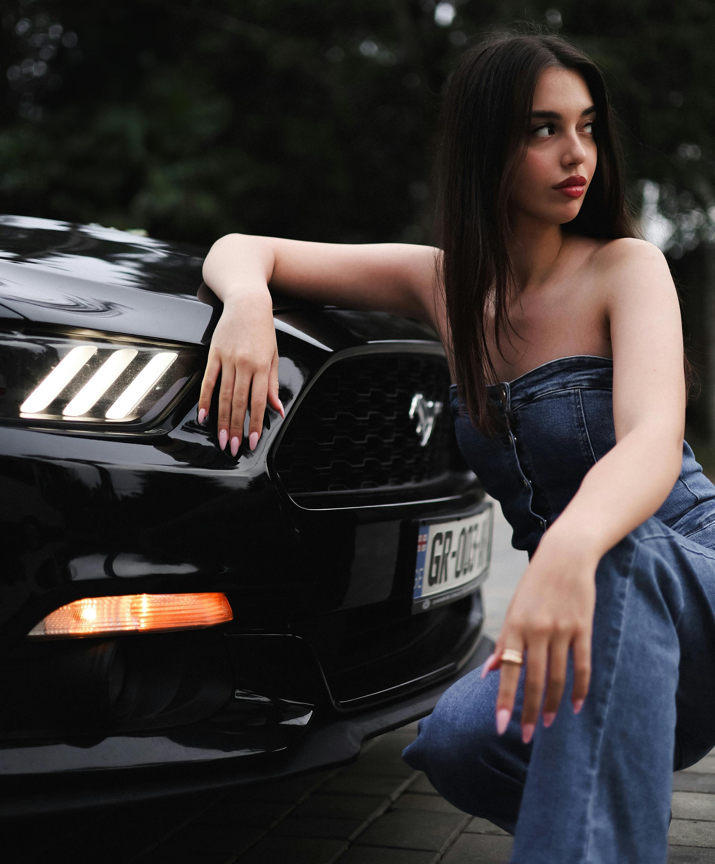 Kostenlos Junge Frau in Jeans posiert neben einem schwarzen Mustang und strahlt Selbstbewusstsein und Stil aus. Stock-Foto