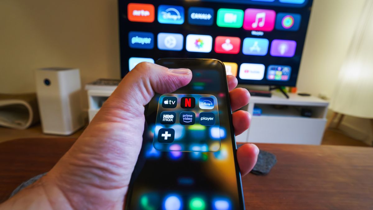 Mão segurando um celular com ícones de aplicativos de streaming em frente a uma televisão